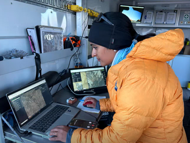 NASA team member in a cold-weather field station reviewing AEGIS traverse maps on a laptop and second monitor