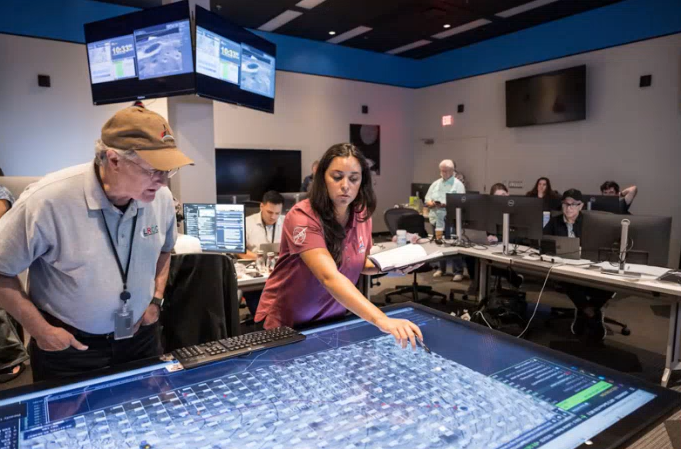 NASA flight controllers reviewing an AEGIS geospatial map on a large touch table in a mission control room